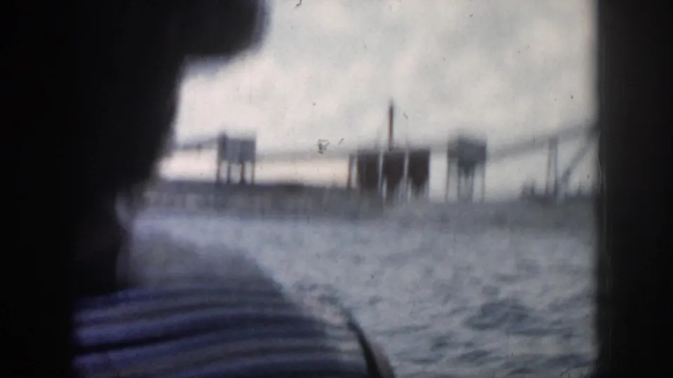 A boat passenger in a striped shirt looks out toward a blurred industrial waterfront in 1958 New Orleans home-movie footage.