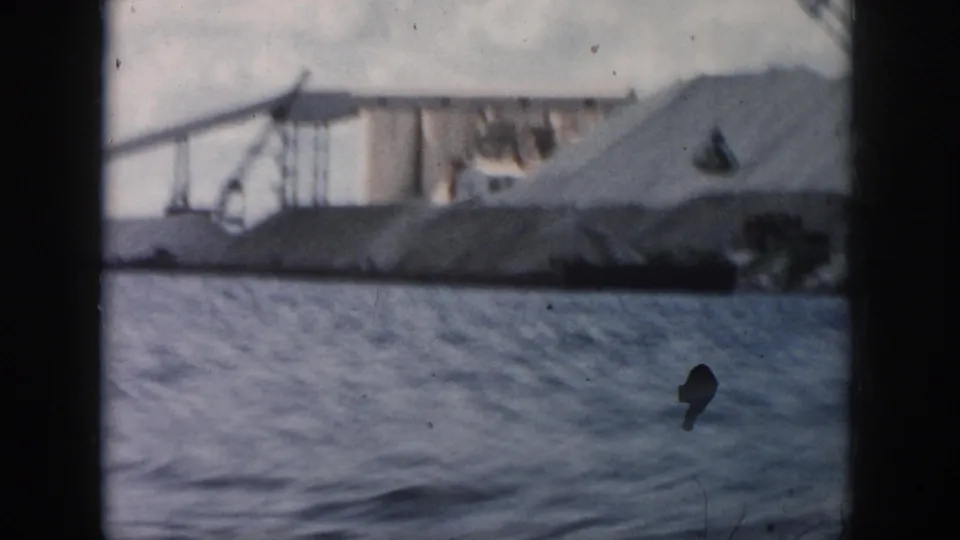 Cranes and pale industrial waterfront forms appear beyond rippling water in the New Orleans boat footage.