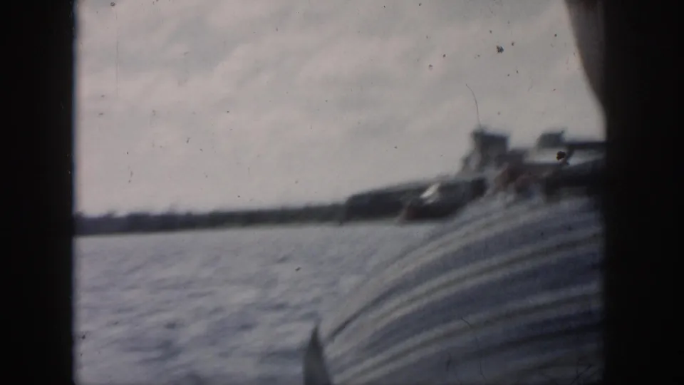 Soft 8mm frame from the moving boat, with a striped passenger in the foreground and distant New Orleans shoreline across the water.