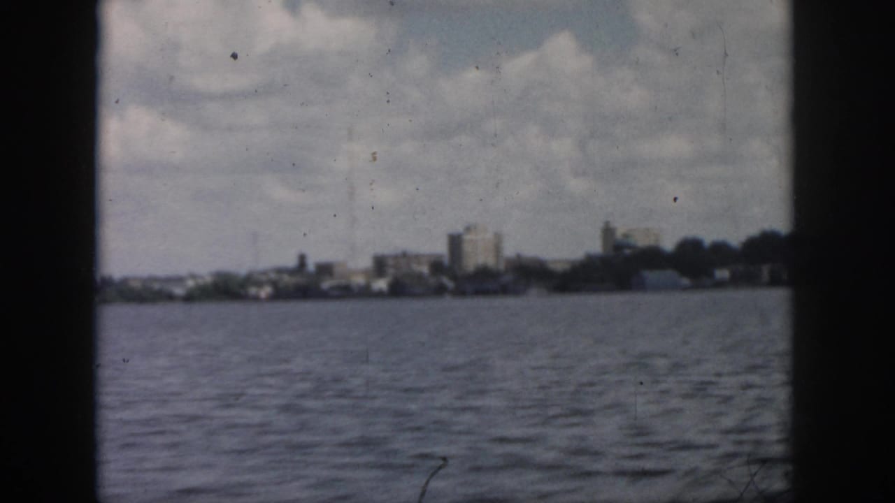 1958 New Orleans waterfront view from across open water, with low buildings, trees, cloudy sky, and dark frame edges.
