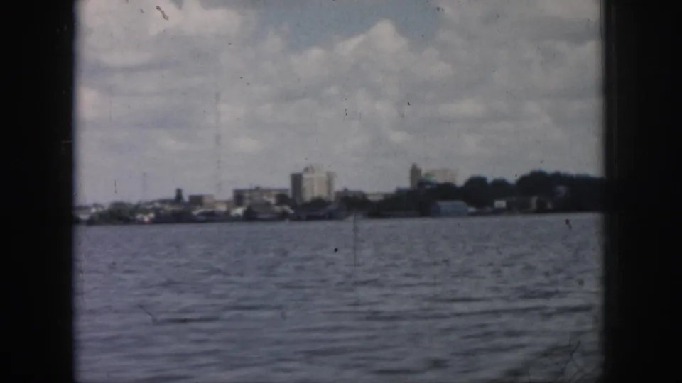 Distant New Orleans shoreline seen from a moving boat, with low-rise buildings, trees, clouds, and 8mm film wear.