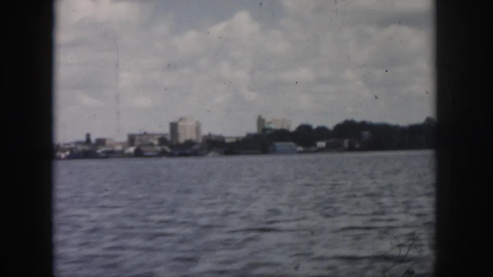 Mid-century 8mm view of a New Orleans waterfront from the water, showing distant buildings and trees beneath clouds.