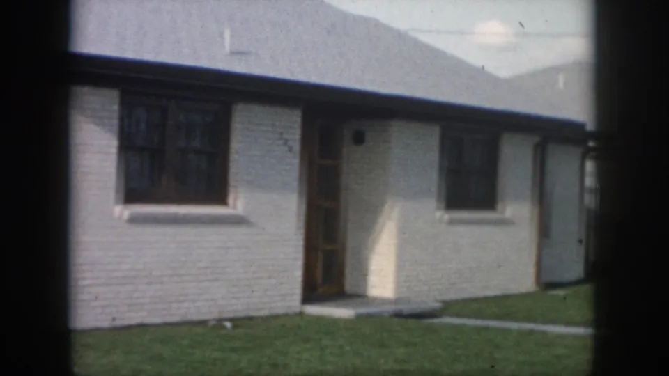 1958 New Orleans exterior view of a white brick ranch house with a wooden front door, dark window trim, and grass lawn.