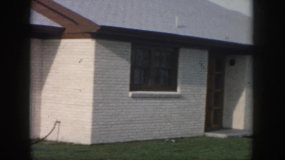 1958 New Orleans home-movie frame showing the house facade, multi-pane window, wooden door, white brick, and green lawn.