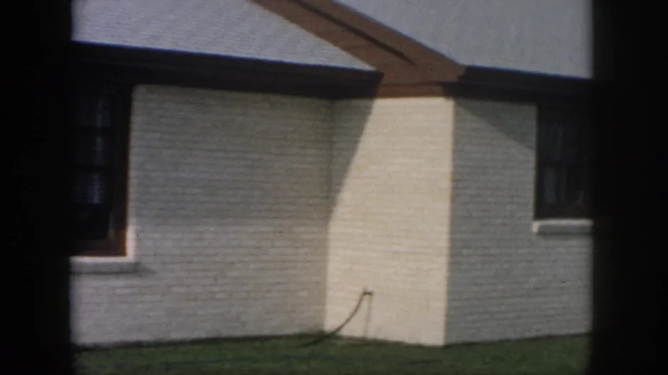 1958 New Orleans frame showing the side wall of a white brick ranch house, green lawn, roofline, and partial window.