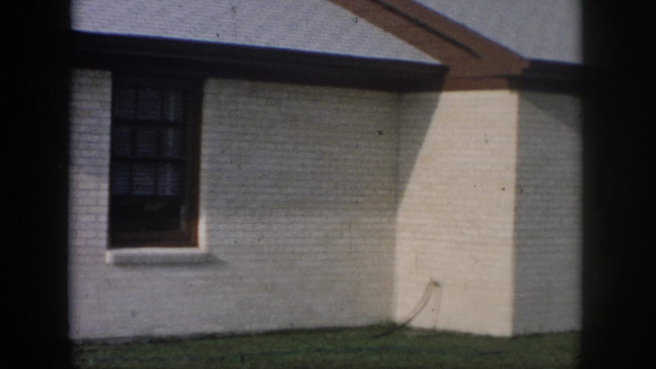 1958 New Orleans home-movie frame showing a white brick residential wall, dark window, grass edge, and roof overhang.