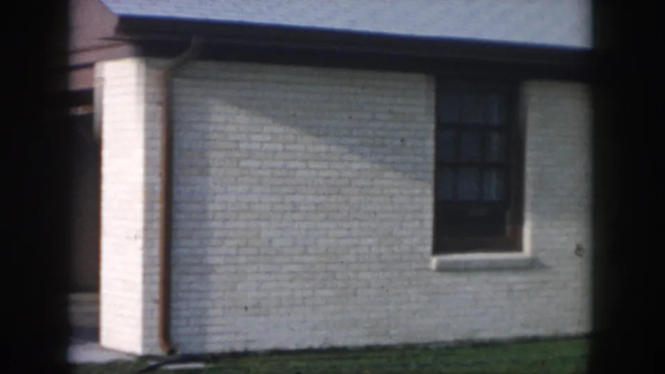 1958 New Orleans home-movie view of a white brick side wall with dark window, gutter downspout, grass, and brown trim.