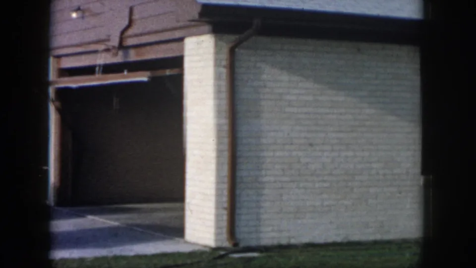 1958 New Orleans residential exterior showing a white brick corner, open garage bay, driveway, gutter, and grass edge.