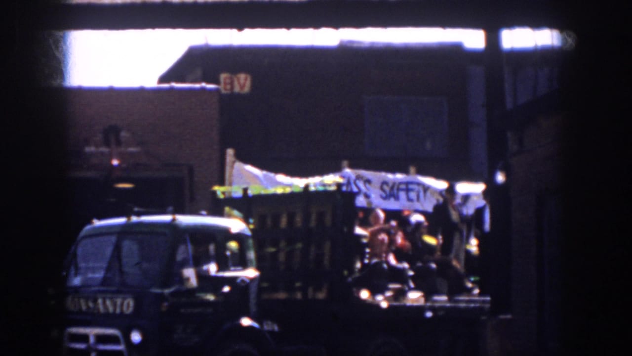 The parade float passes through the frame with a visible Monsanto truck cab, costumed riders, and a partially readable white banner.