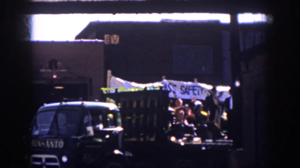 The parade float passes through the frame with a visible Monsanto truck cab, costumed riders, and a partially readable white banner.