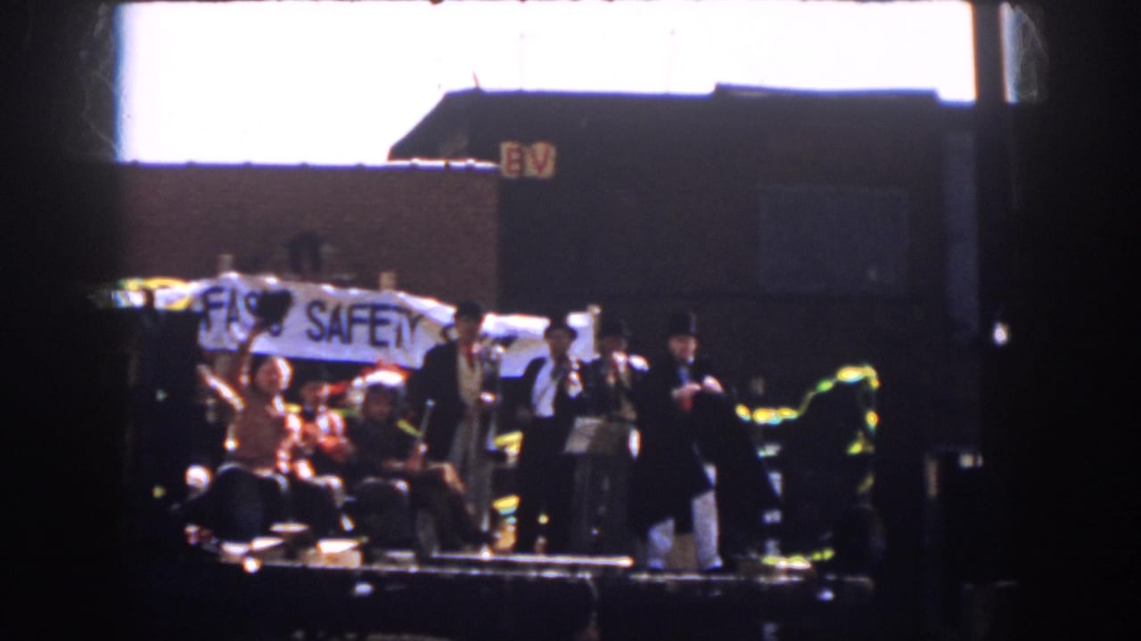 1958 Virginia parade view with suited men and seated riders on a flatbed trailer carrying a safety banner.