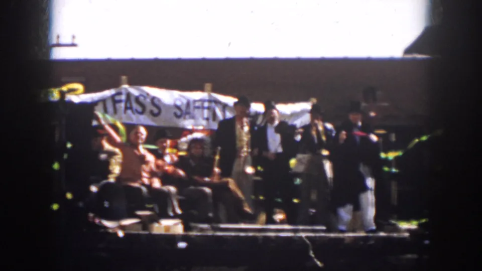 1958 Virginia parade frame with people on a flatbed trailer, dark-suited men, and a large FAS'S SAFETY banner.