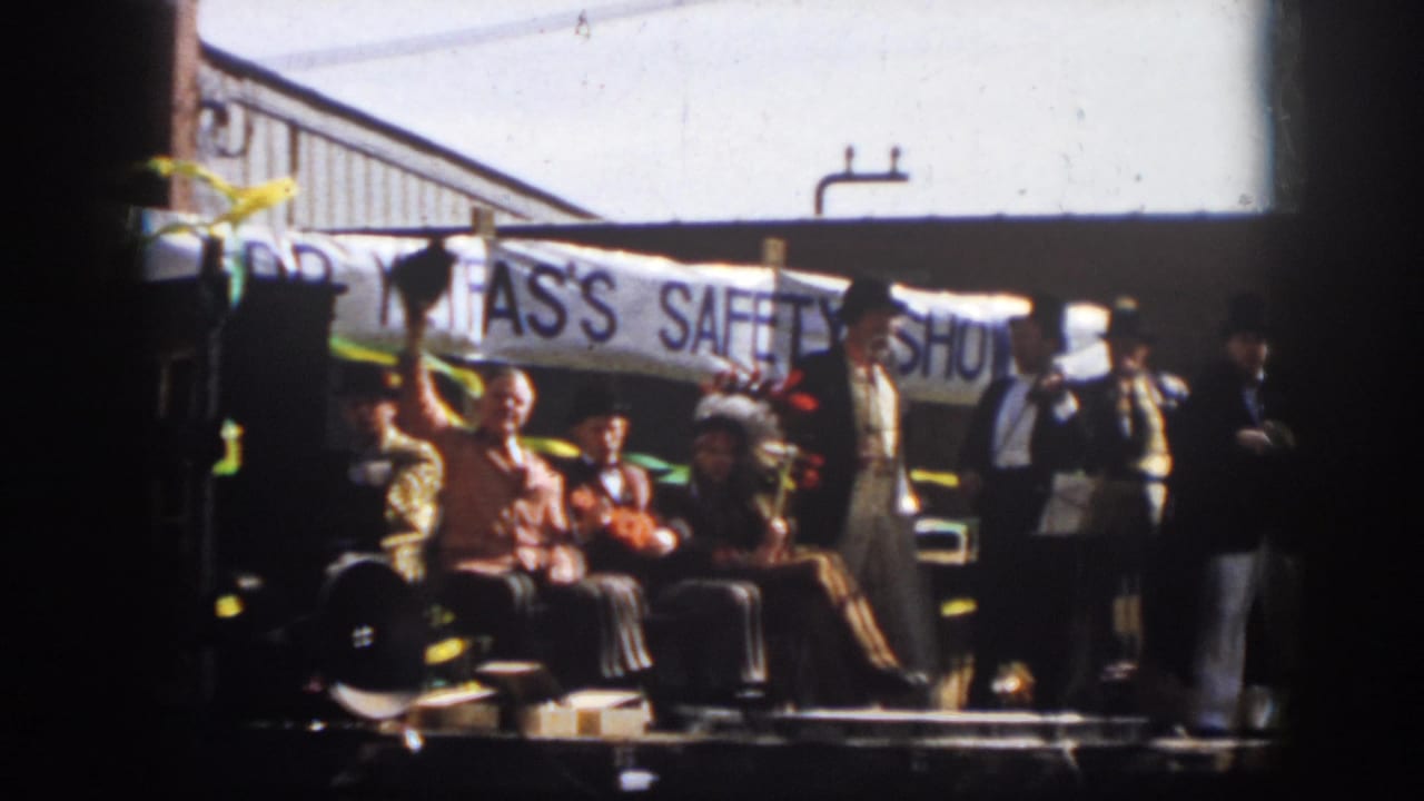 Costumed men on a parade float sit in front of a banner while another man stands at the side.