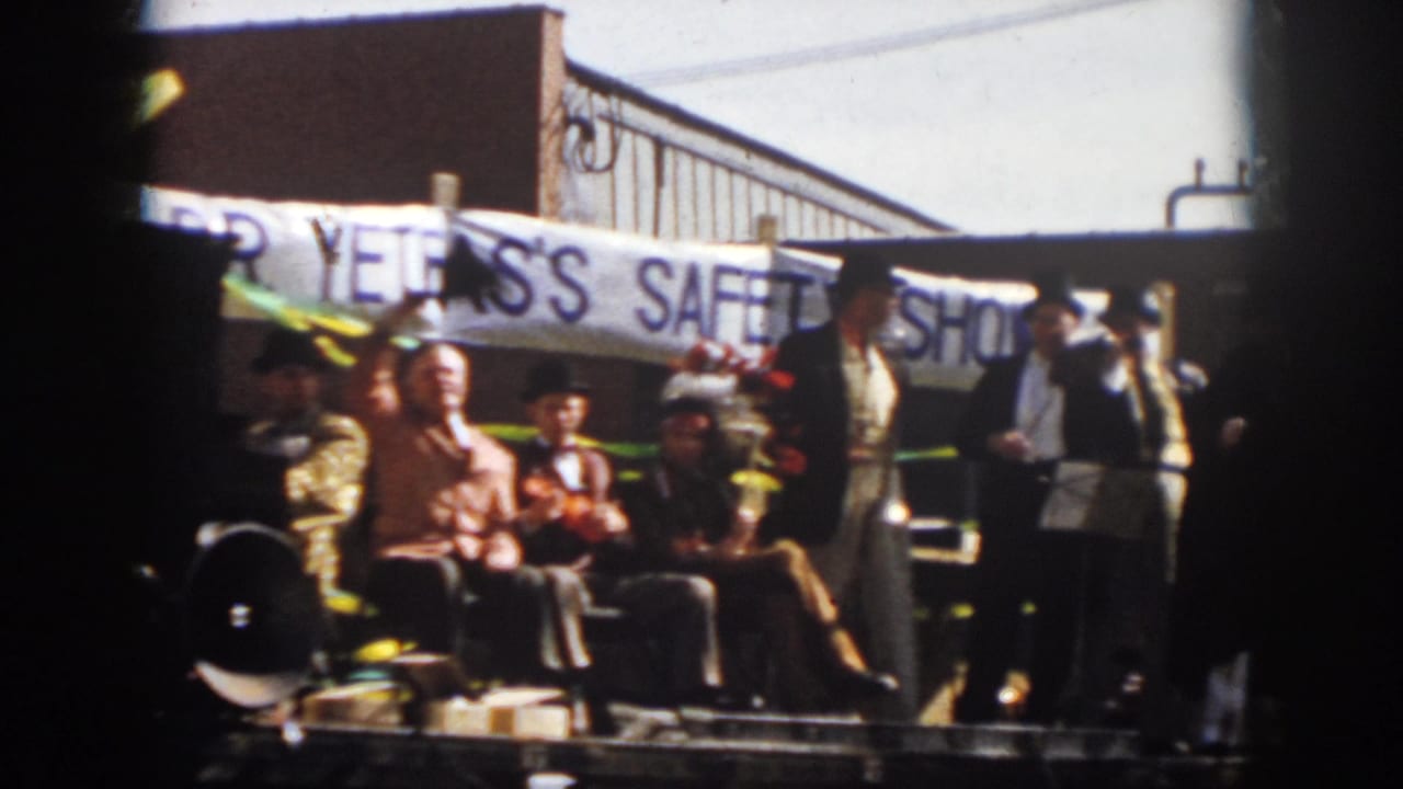 1958 Virginia home-movie frame of a parade float with seated men, formal costumes, headdress, and a safety-show banner.