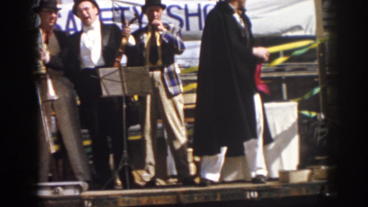 Men in formal clothing stand near trombones, music stands, and a decorated float backdrop.