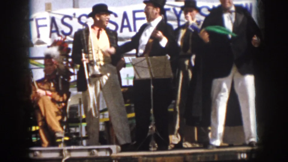 1958 Virginia home-movie frame of performers pausing on a parade float, with trombone, music stand, and seated players visible.