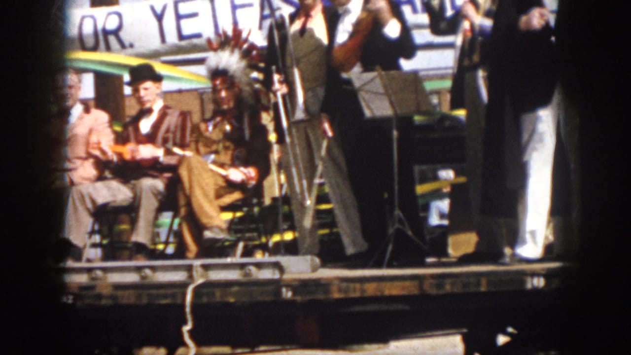Seated performers on a float with instruments, headdress, microphone stands, and a partial banner behind them.
