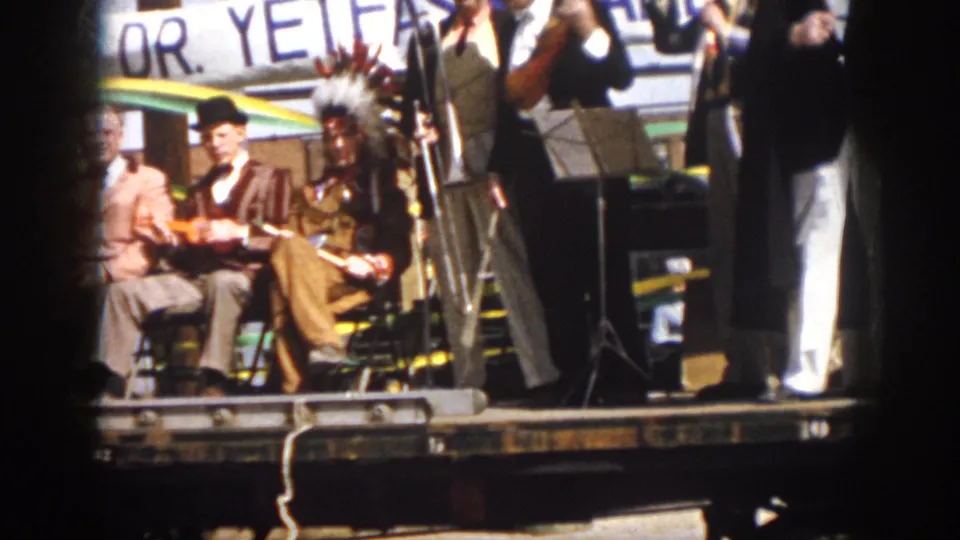 Virginia 1958 parade float scene with seated musicians, a headdressed performer, microphone stands, and a partially readable background banner.