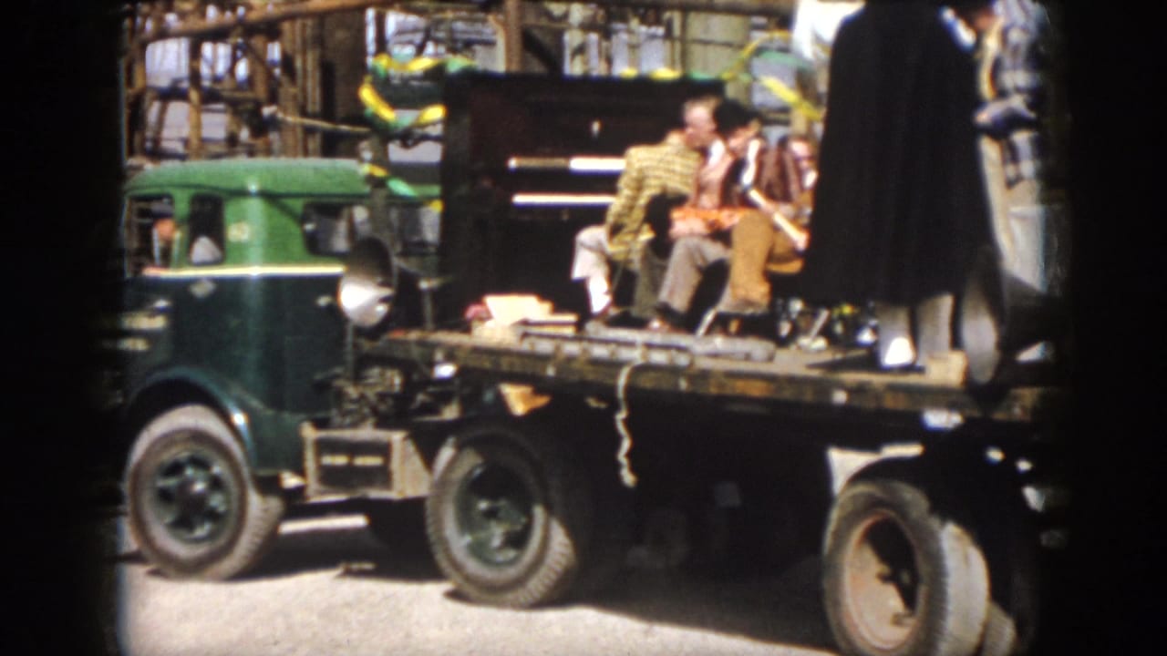 Wider 1958 Virginia home-movie view of a green truck pulling a flatbed float with performers, instruments, and street parade equipment.