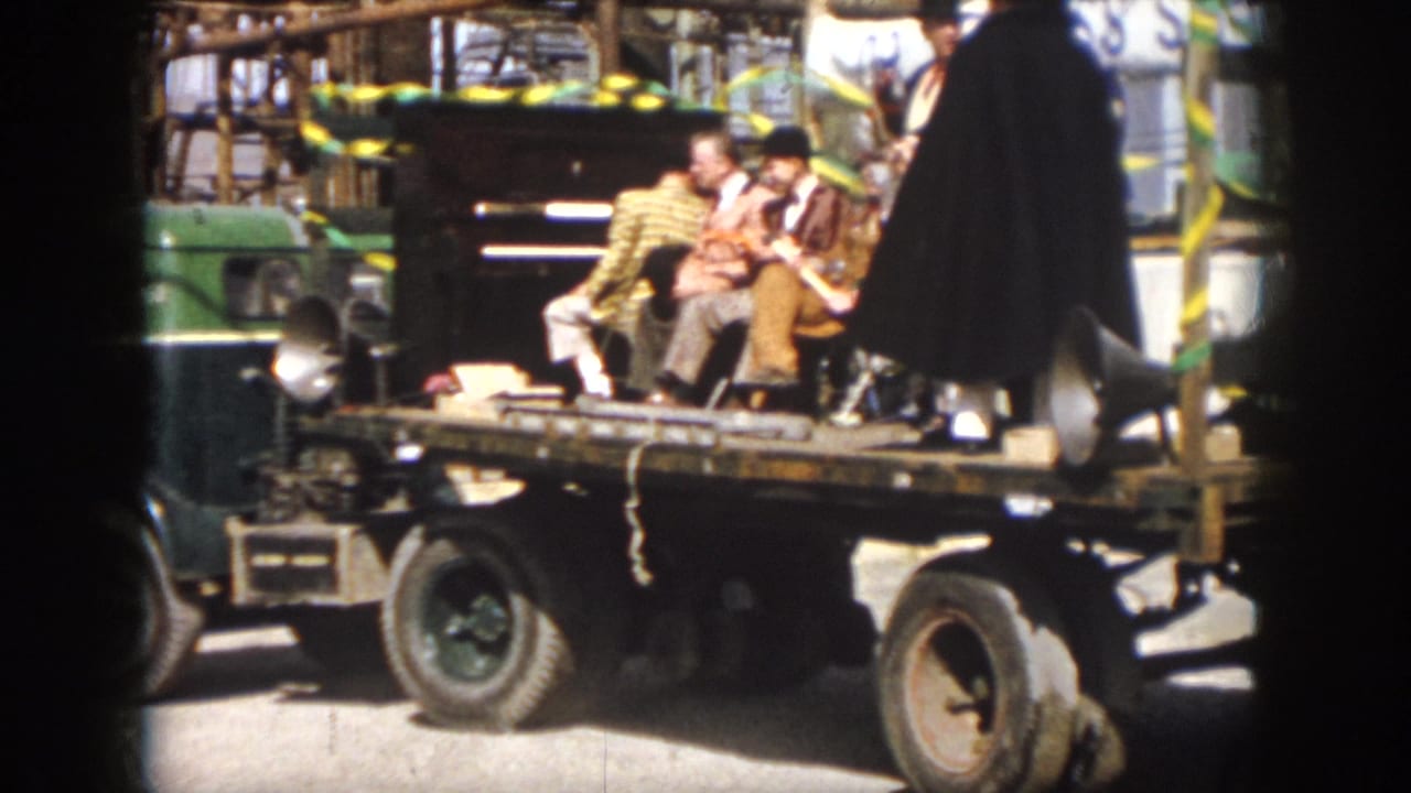 1958 Virginia home-movie frame showing a decorated flatbed float with seated men, ribbons, a loudspeaker, and a caped performer standing nearby.