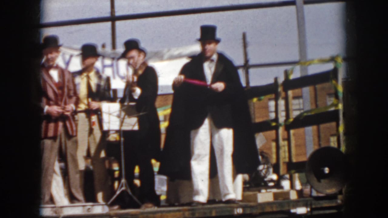 Magician and suited performers stand on a flatbed truck stage before workers in an industrial yard.