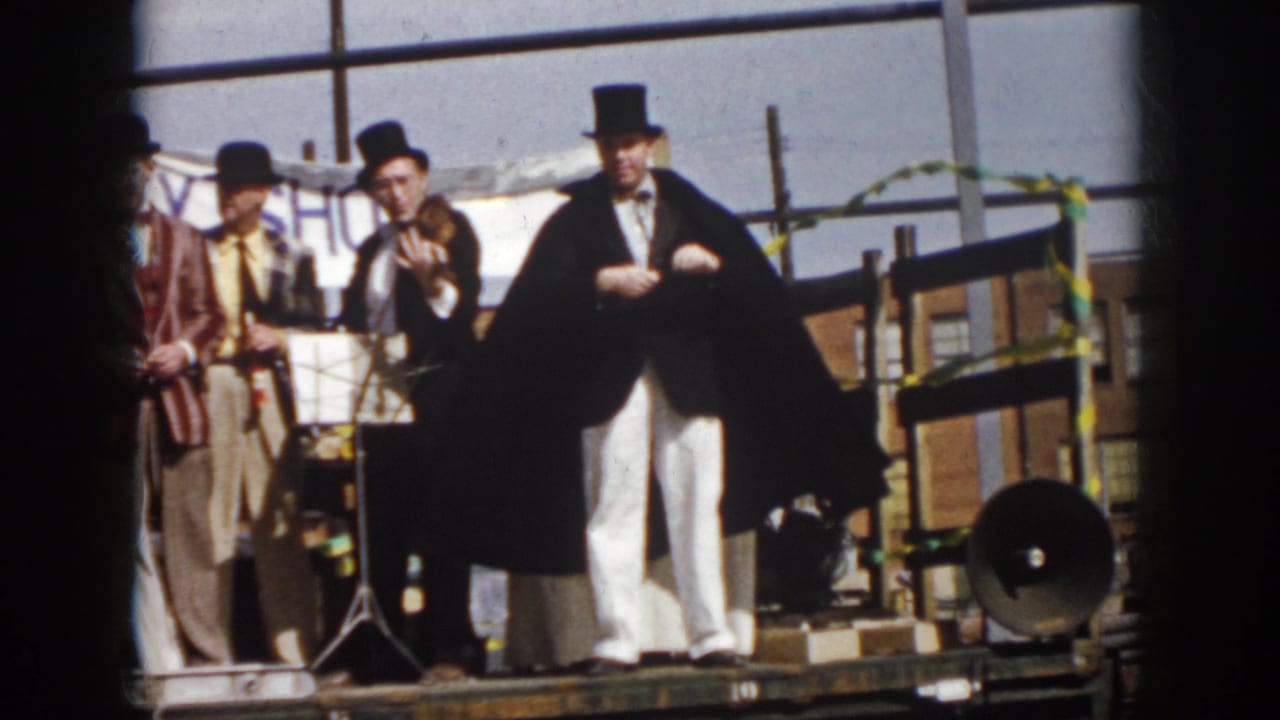 1958 Virginia frame of a magician in top hat and cape performing beside men in hats on a flatbed truck stage.