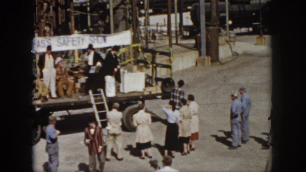 1958 Virginia home-movie frame showing spectators around a flatbed truck stage with a safety show banner and industrial pipes behind it.