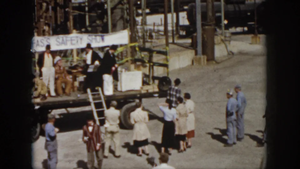 1958 Virginia home-movie frame showing spectators around a flatbed truck stage with a safety show banner and industrial pipes behind it.
