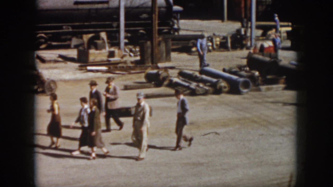 In 1958 Virginia footage, adults in suits, hats, and dresses walk through a railroad yard near rail tank cars, pipes, and workers.