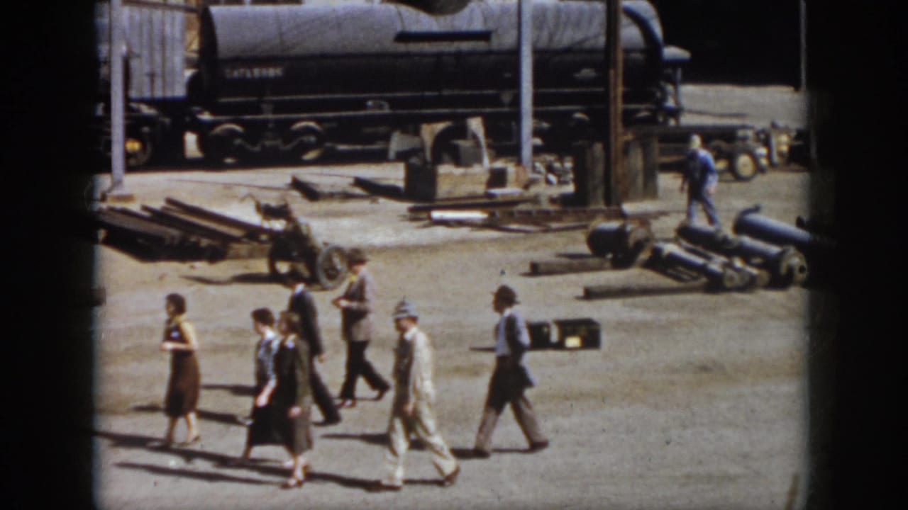 Adults continue walking past rail-yard pipes, poles, and a dark tank car in the background.