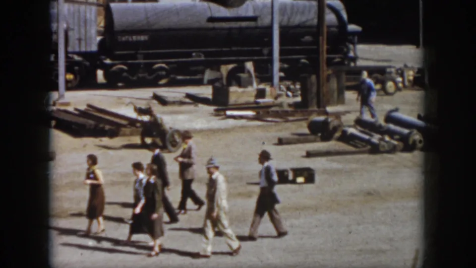 Adults in mid-century clothing walk across a Virginia railroad yard with a tank car, scattered industrial materials, and workers visible behind them.