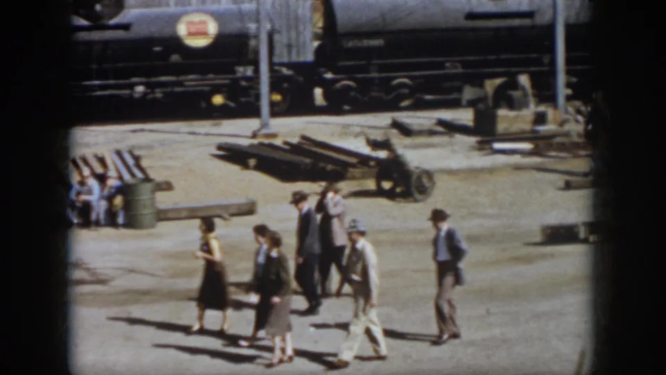 The 1958 Virginia home movie shows adults walking through a rail yard while workers sit near barrels and materials below a dark tank car.