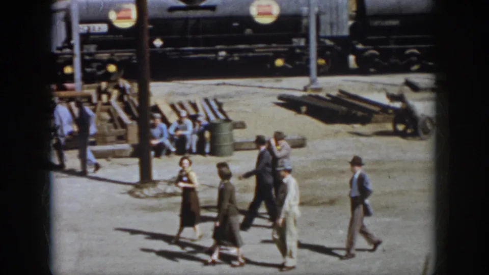 A group in 1950s clothing walks through a Virginia railroad yard past workers, barrels, stacked rails, poles, and a dark tank car.