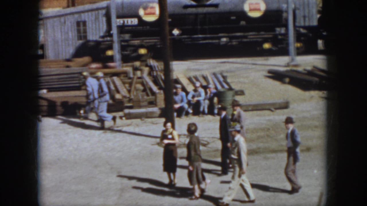 Virginia 1958 home-movie frame showing workers and visitors in an industrial yard with stacked lumber or pallets and a parked vehicle nearby.