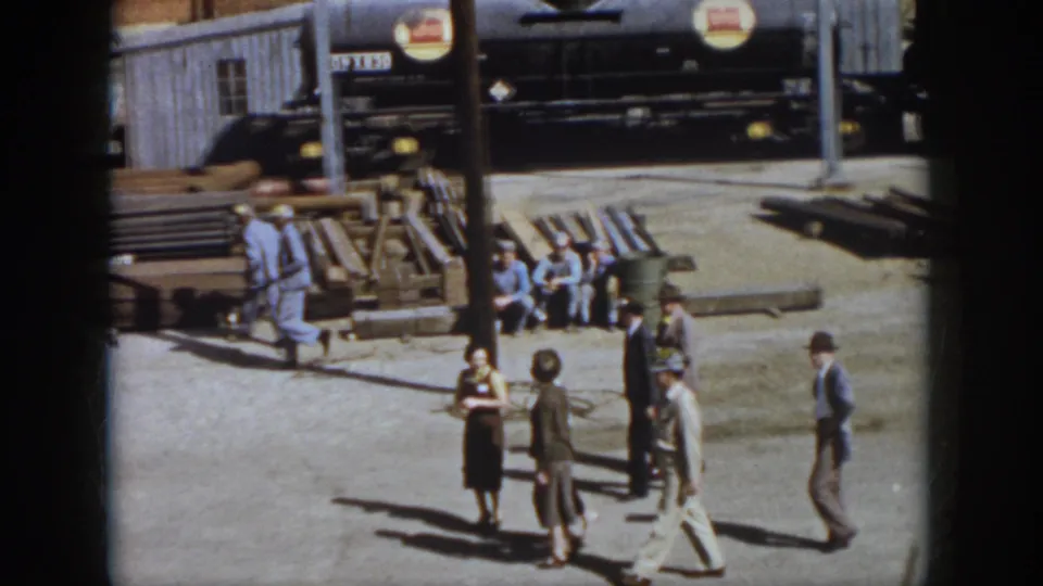 Virginia 1958 home-movie frame showing workers and visitors in an industrial yard with stacked lumber or pallets and a parked vehicle nearby.