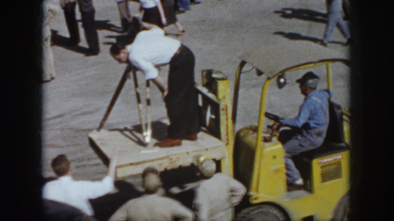 Virginia 1958 frame with a man using crutches on a wooden platform next to a yellow forklift while workers watch in the yard.