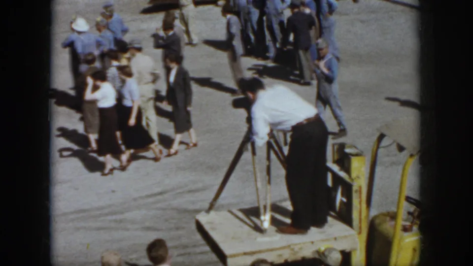 Virginia 1958 frame showing visitors and workers watching a man with crutches on a platform next to a yellow forklift in bright sun.