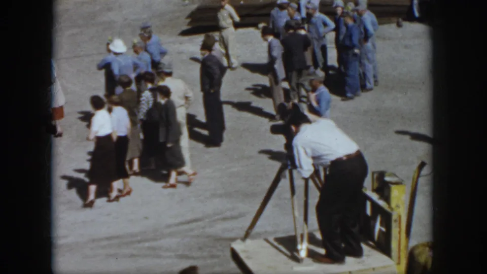 1958 Virginia home-movie frame showing adults gathered on pavement while a man in a white shirt films from a tripod camera.