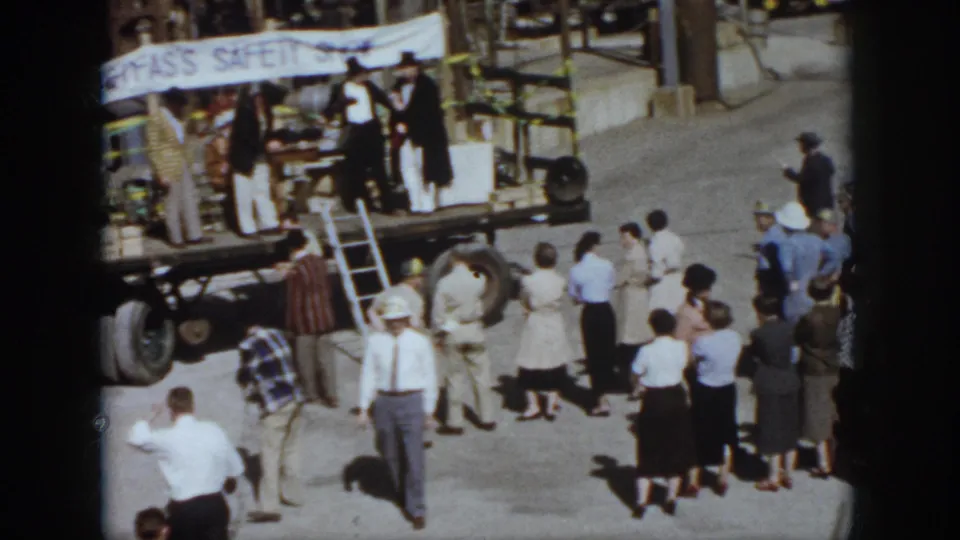 1958 Virginia event frame showing spectators facing a small raised platform or trailer with a partly readable safety banner.