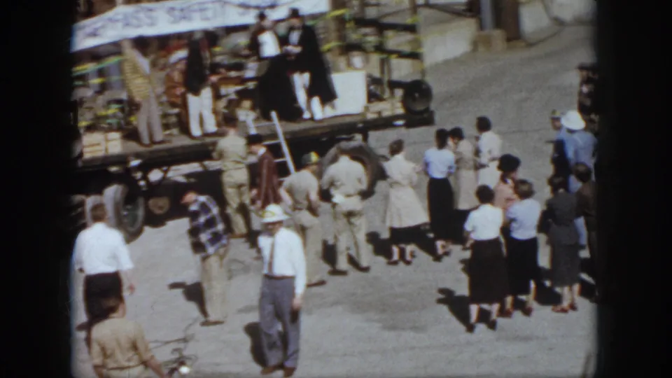 1958 Virginia home-movie frame of adults gathered near a small event platform or trailer with a partial safety banner overhead.