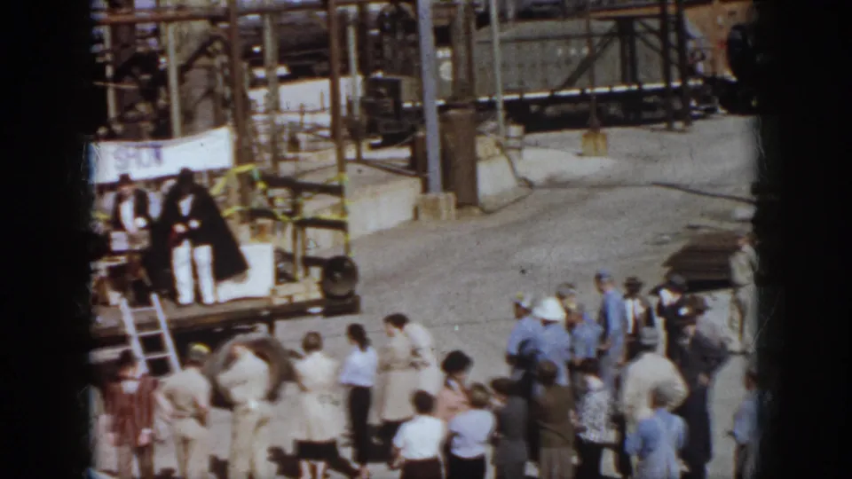 A 1958 Virginia crowd stands in a railway yard facing a safety-show platform, with freight cars and pipework in the background.