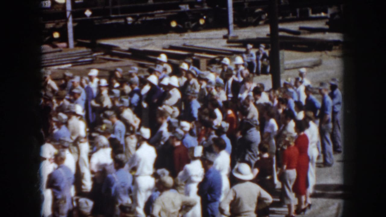 1958 Virginia frame of adults in mid-century clothing gathered at a train yard, with freight cars and track materials visible behind them.