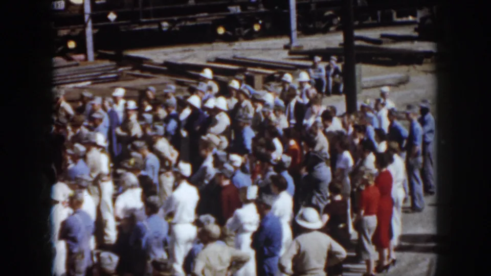 1958 Virginia frame of adults in mid-century clothing gathered at a train yard, with freight cars and track materials visible behind them.