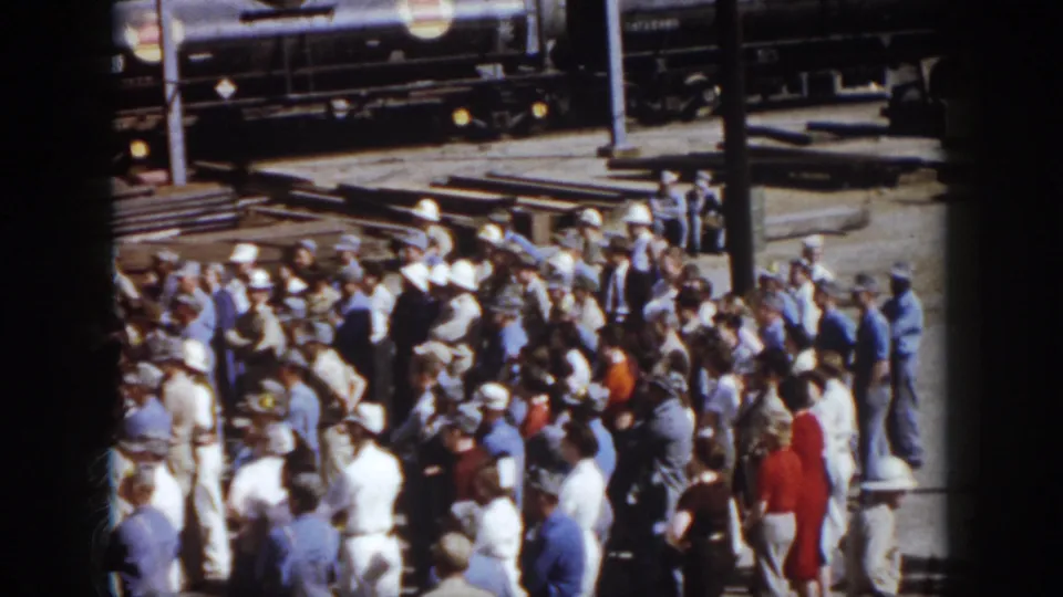 Elevated 1958 Virginia home-movie view of adults packed near a rail yard, with track hardware, freight cars, and bright clothing visible.