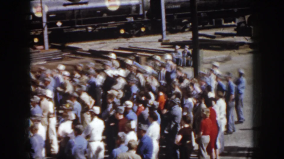 Late shot from the 1958 Virginia clip showing a dense adult crowd beside rail tracks, freight cars, and strong sunlight on aged 8mm film.