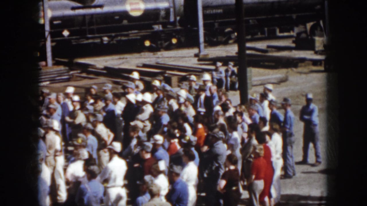 1958 Virginia home-movie frame showing a large outdoor crowd near rail cars and factory structures at an industrial site.