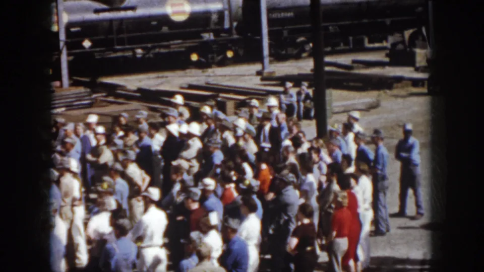 1958 Virginia home-movie frame showing a large outdoor crowd near rail cars and factory structures at an industrial site.