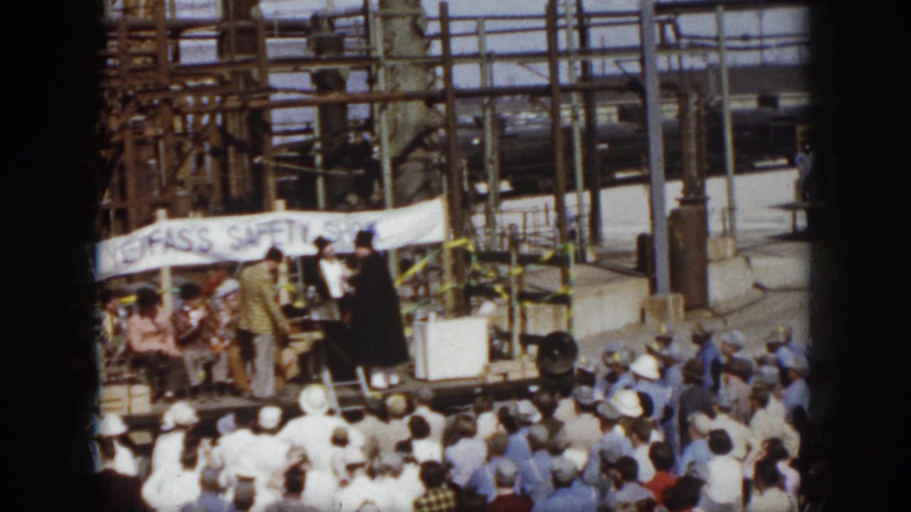 Outdoor stage with speakers, seated people, banner, and crowd below.
