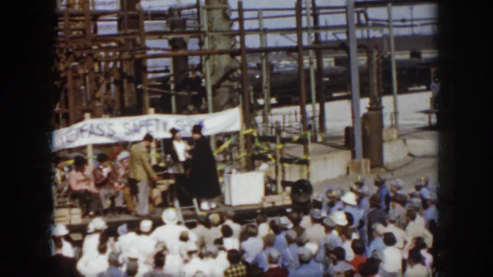 1958 Virginia frame showing a makeshift stage at an industrial site, with a safety-show banner and crowd watching.