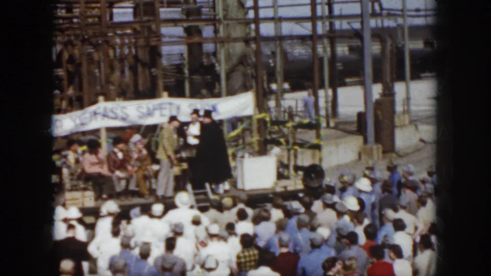 1958 Virginia home-movie view of a crowd watching a small stage backed by factory structures and a safety-show banner.